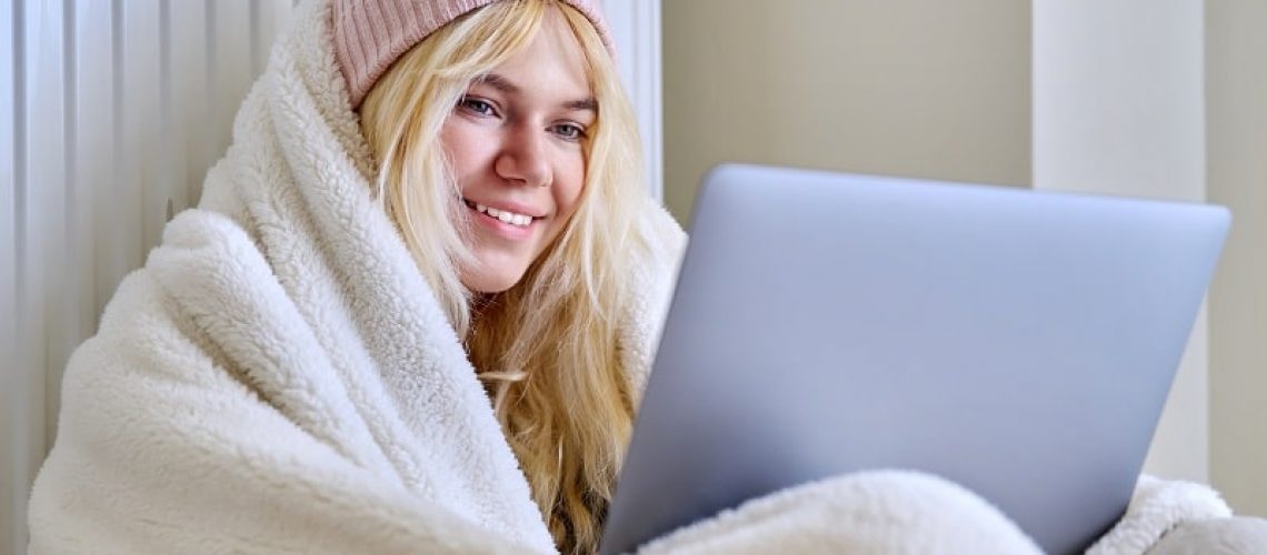 A young female teenager frozen in house in winter cold season, warming up with a warm blanket, hat, central heating radiator, looking at laptop screen, student, having rest