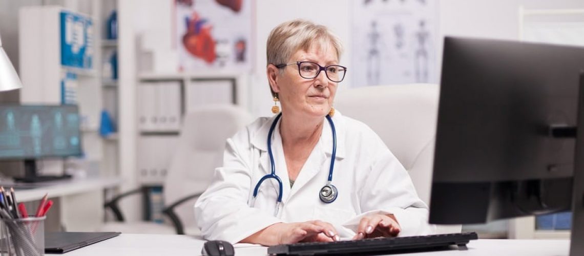 Mature female doctor using computer in hospital cabinet.